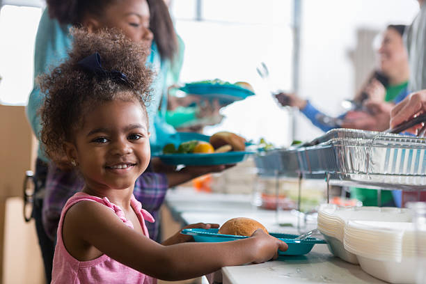 Cheerful African American little girls smiles while in line in a soup kitchen. She is holding a plate full of healthy food. Her family is in line behind her.